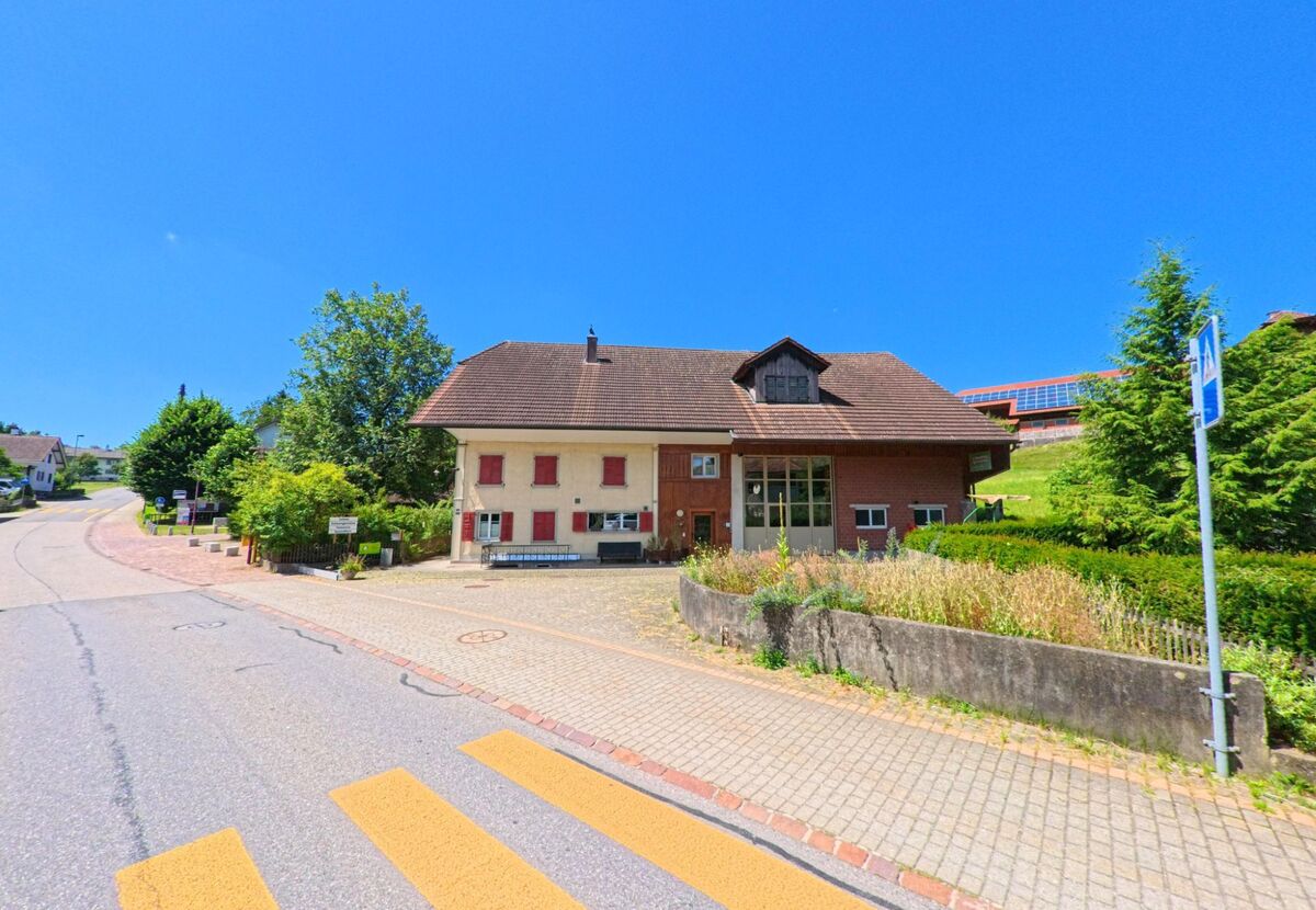 Ein Haus mit roten Fensterlaeden und angeschlossener Halle steht an einem Fussgaengerstreifen in Roggliswil (PLZ 6265, Kanton Luzern), im Hintergrund ist ein mit Solarpanels gedecktes Gebaeude sichtbar.