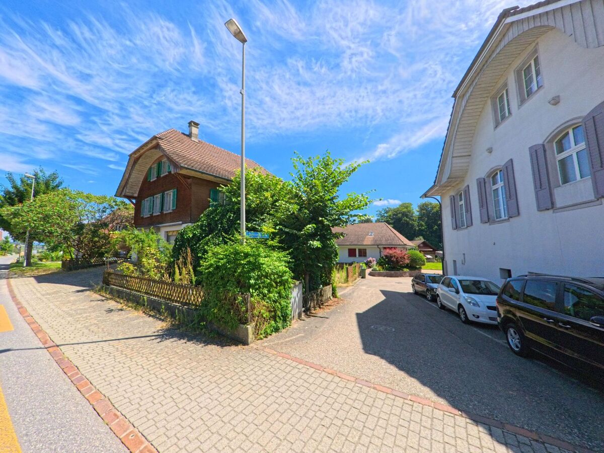 Zwischen zwei Wohnhaeusern in Roggliswil (PLZ 6265, Kanton Luzern) zweigt eine schmale Seitenstrasse ab, die von parkierten Autos und gruener Bepflanzung gesaeumt ist.