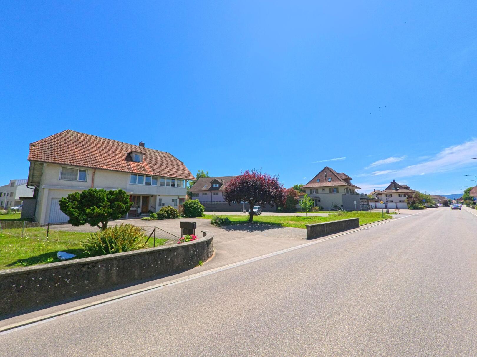 In Roggwil (4914, Kanton Bern) stehen Einfamilienhaeuser mit gepflegten Vorgartenanlagen entlang einer breiten, sonnigen Strasse.
