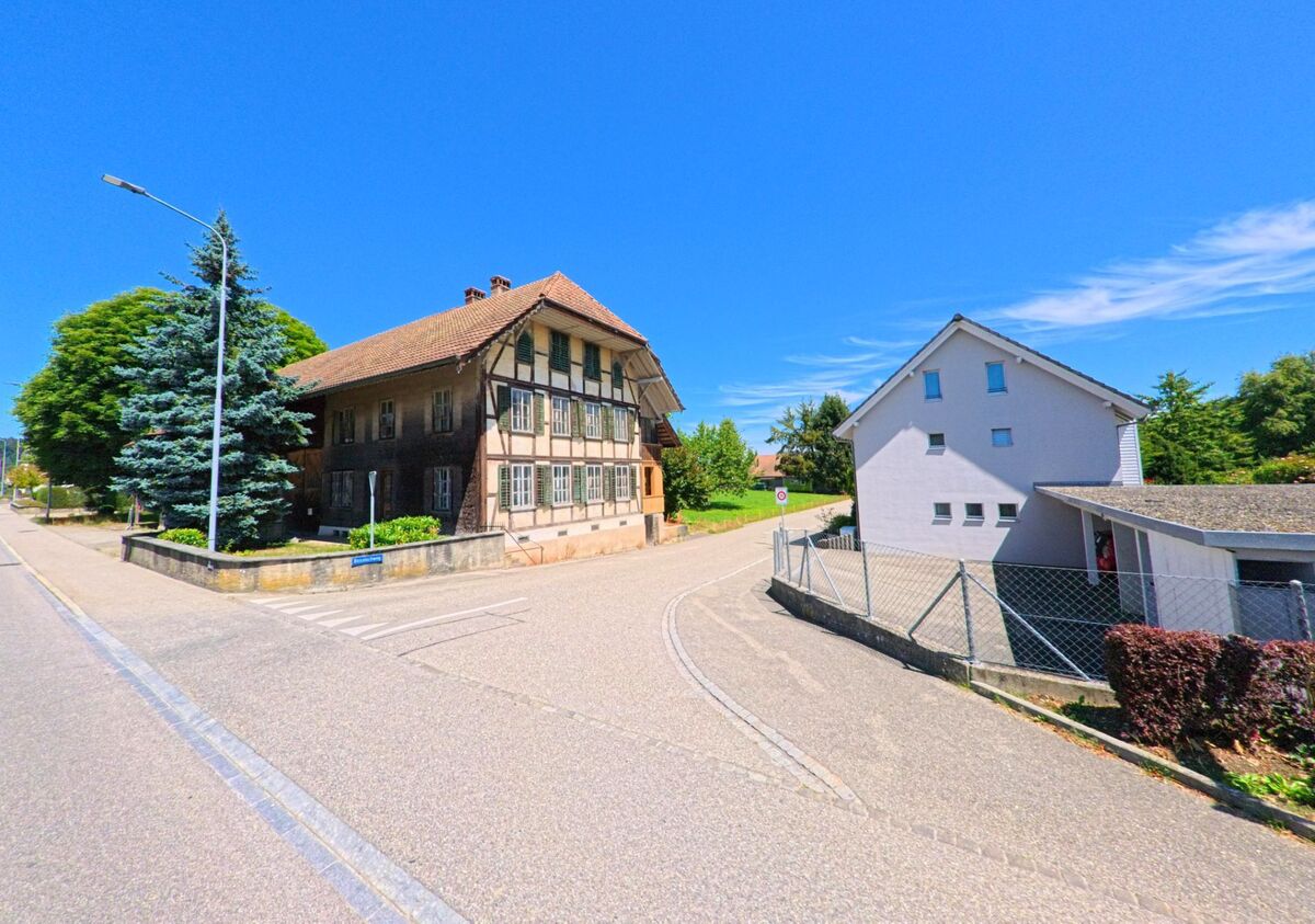 In Roggwil (4914, Kanton Bern) steht ein historisches Fachwerkhaus mit gruenen Fensterlaeden an einer ruhigen Strassenecke neben modernen Wohnbauten.