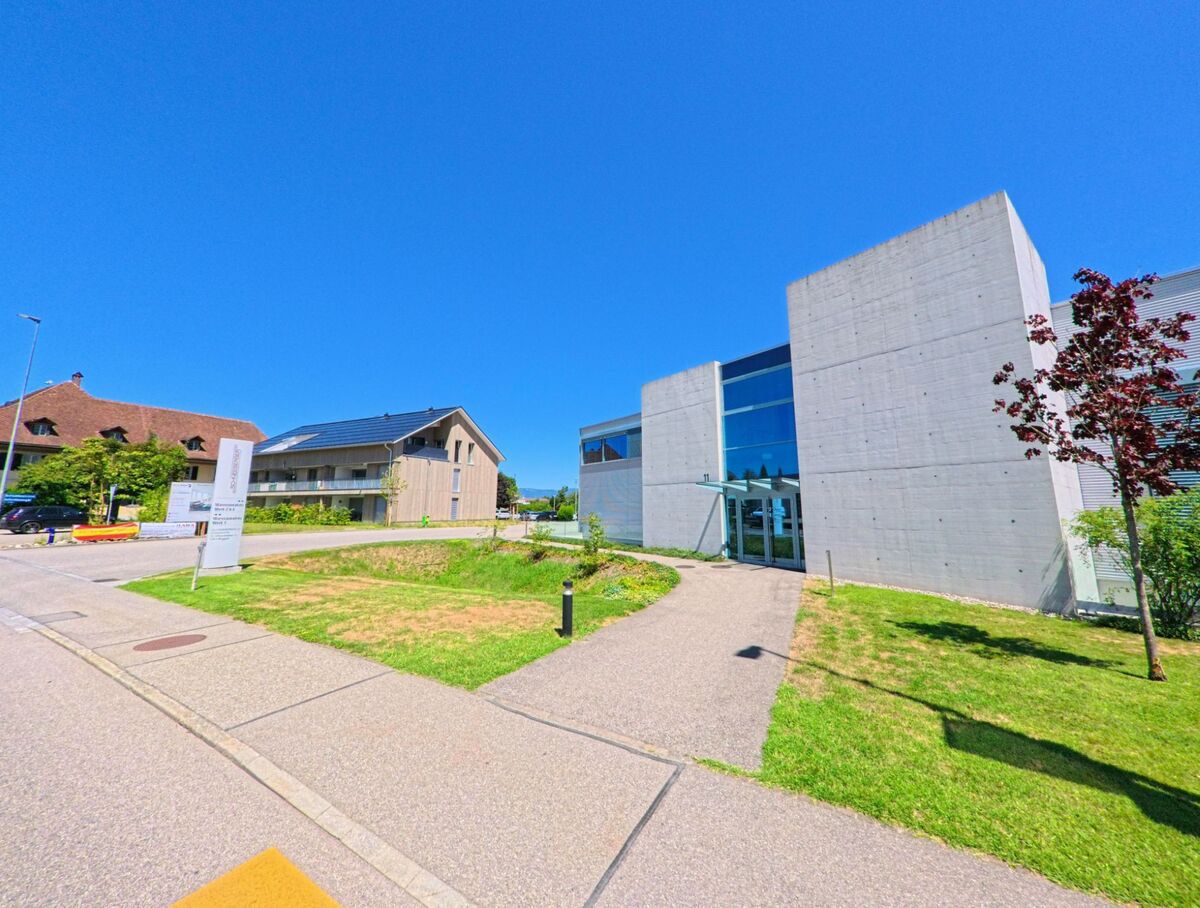 In Roggwil (4914, Kanton Bern) steht ein modernes Gebaeude mit Glasfassade und Sichtbeton neben einer gepflegten Gruenanlage und weiteren zeitgenoessischen Bauten.