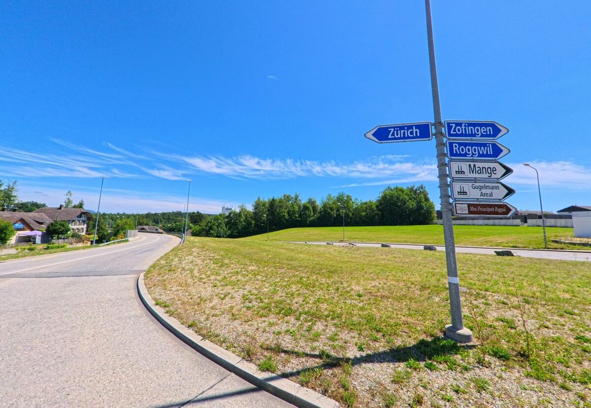 Wegweiser am Ortseingang von Roggwil (4914, Kanton Bern) zeigen Richtungen nach Zuerich, Zofingen und zum Guggelmann Areal vor einer offenen Wiesenlandschaft.