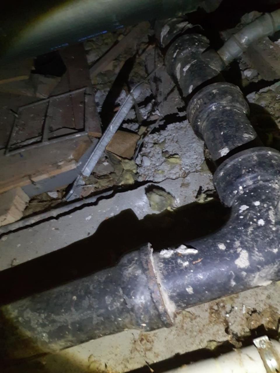 In einem dunklen Raum ist ein beschädigtes Rohr zu sehen, umgeben von Staub und losem Material auf dem Boden.