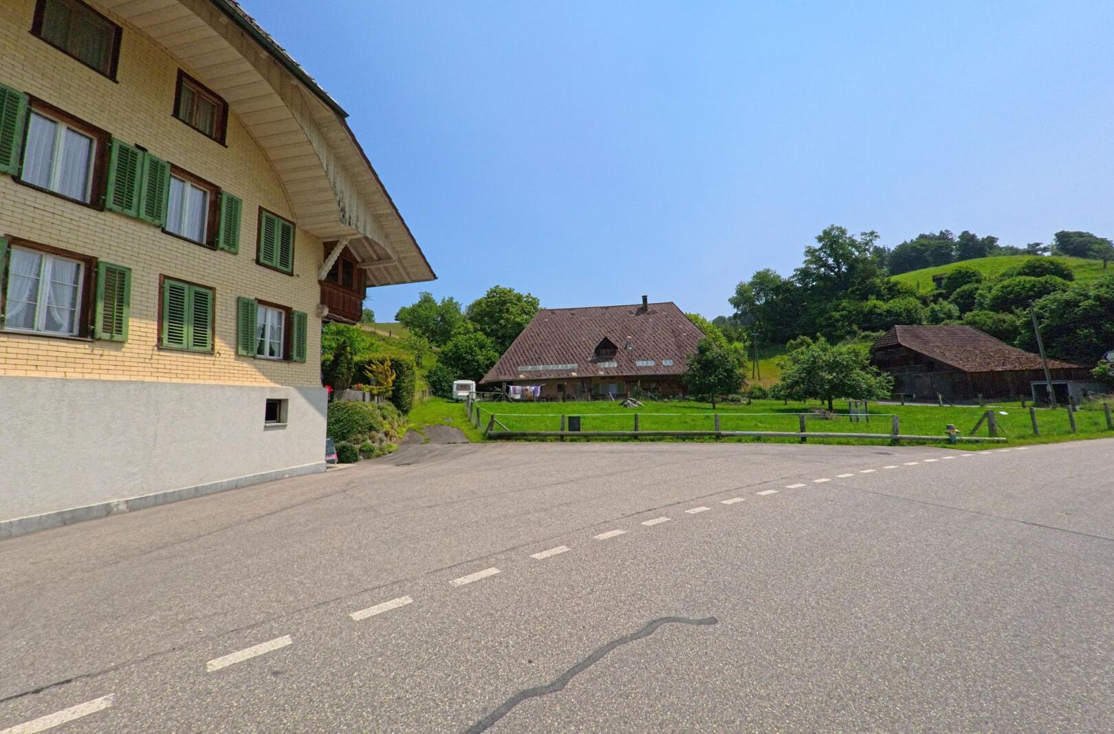 An einer sanft geschwungenen Strasse in Rohrbachgraben (4938, Kanton Bern) stehen traditionelle Bauernhäuser mit grünen Fensterläden vor einer grünen Hügellandschaft.