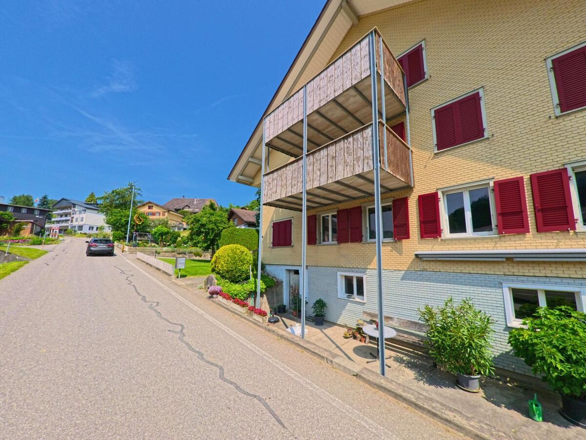 In Rohrbachgraben (4938, Kanton Bern) säumt ein gelbes Mehrfamilienhaus mit roten Fensterläden und Holzverkleidungen einen leicht ansteigenden Wohnstrassenabschnitt.