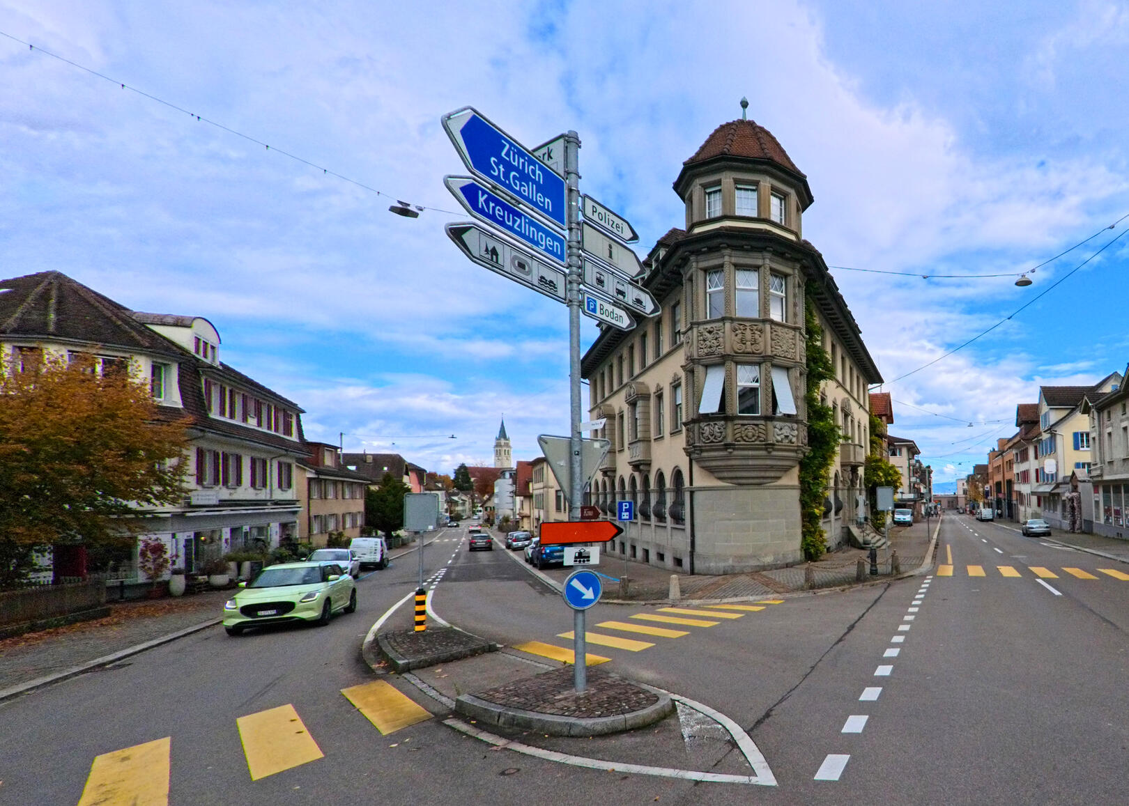 An einer Kreuzung in Romanshorn (8590, Kanton Thurgau) stehen mehrere Wegweiser neben einem markanten Eckgebaeude, waehrend Autos und Fussgaengerstreifen die Strasse strukturieren.