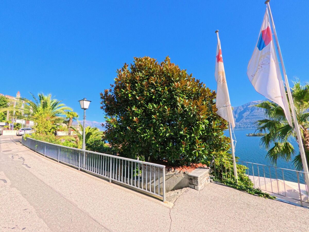 In Ronco sopra Ascona (6613, Kanton Tessin) führt eine steile Strasse mit Geländer und Palmen vorbei an zwei wehenden Fahnen zum Lago Maggiore.