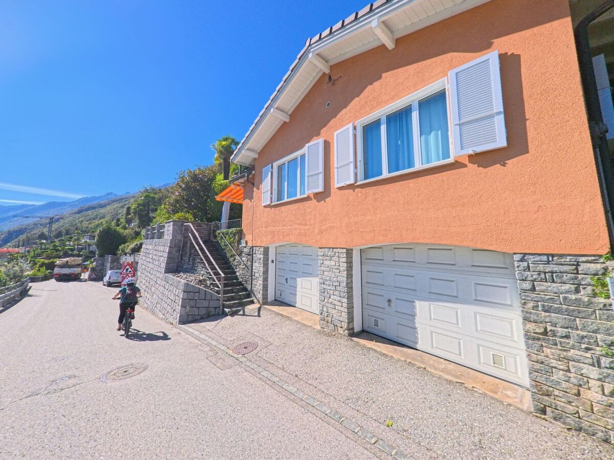 In Ronco sopra Ascona (6622, Kanton Tessin) fährt ein Kind mit dem Fahrrad eine steile Strasse hinab, vorbei an einem orangefarbenen Haus mit Garagen.