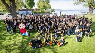 Auf einer Gruenflaeche nahe dem See versammelt sich in Arbon (9320, Kanton Thurgau) eine grosse Gruppe von Kindern, Jugendlichen und Erwachsenen mit Musikinstrumenten zu einem gemeinsamen Foto.