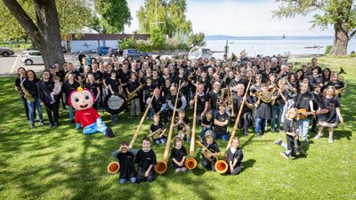 Auf einer Gruenflaeche nahe dem See versammelt sich in Arbon (9320, Kanton Thurgau) eine grosse Gruppe von Kindern, Jugendlichen und Erwachsenen mit Musikinstrumenten zu einem gemeinsamen Foto.