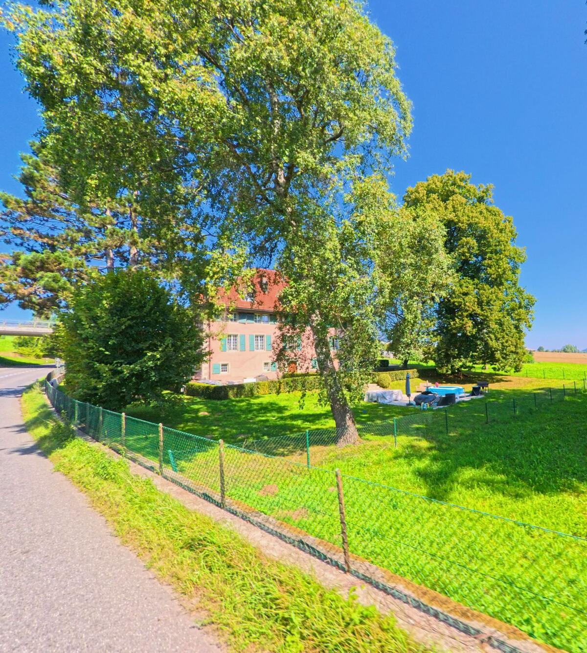 Hinter grossen Baeumen steht in Ropraz (1088, Kanton Waadt) ein mehrgeschossiges Haus mit roten Dachziegeln.