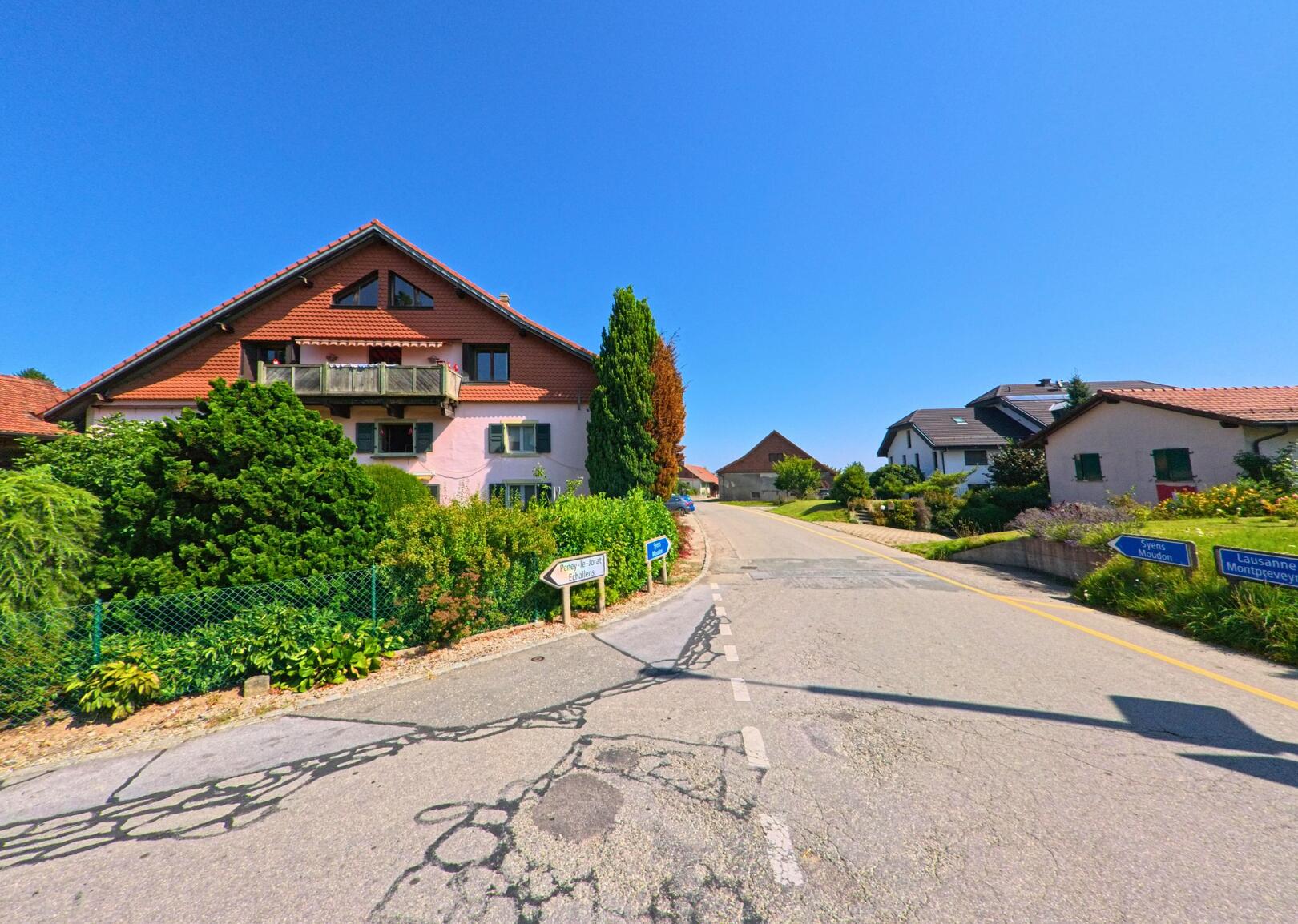 In Ropraz (1088, Kanton Waadt) fuehrt eine Strassenkreuzung an Wohnhaeusern mit roten Daechern vorbei, flankiert von Wegweisern nach Lausanne und Moudon.