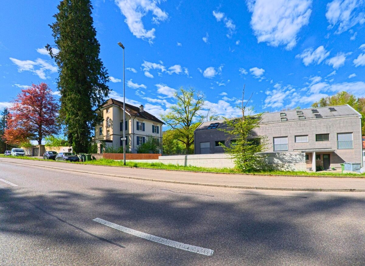 In Rorbas im Kanton Zürich, PLZ 8427, stehen ein historisches Wohnhaus und ein modernes Gebäude nebeneinander an einer leicht ansteigenden Strasse, eingerahmt von hohen Bäumen.