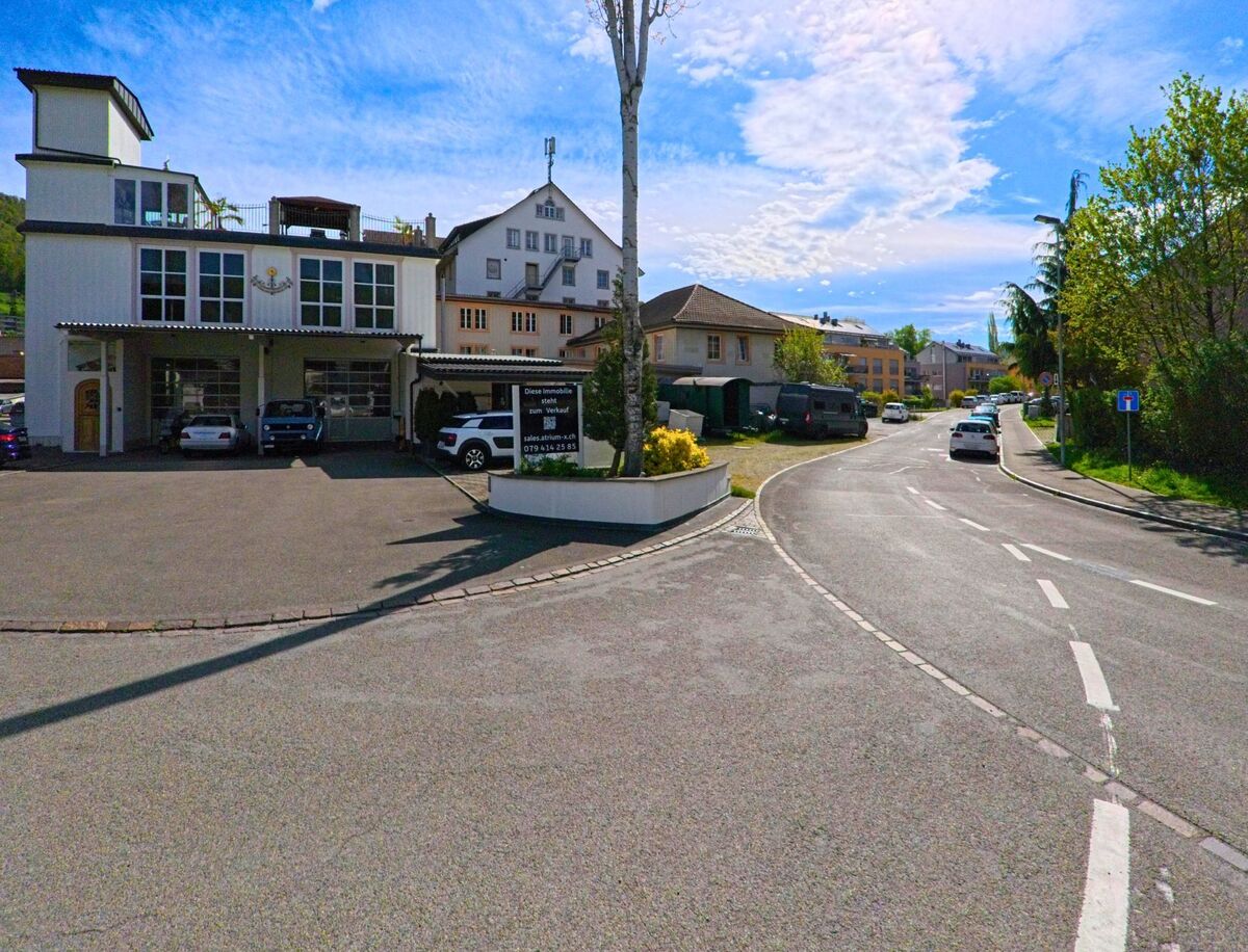 Im Ort Rorbas, PLZ 8427 im Kanton Zürich, verläuft eine leicht geschwungene Strasse an einem modernen Gewerbegebäude mit mehreren geparkten Fahrzeugen vorbei.