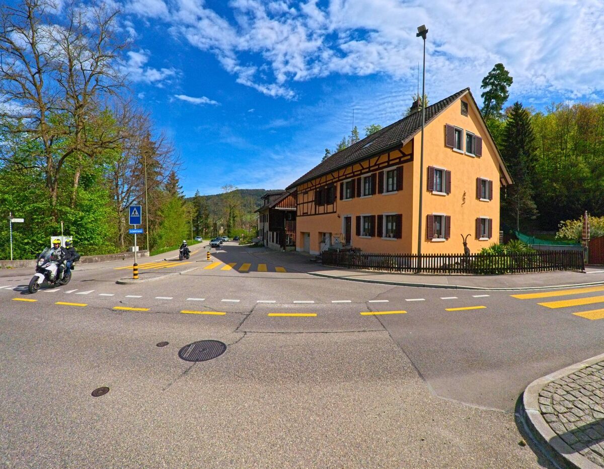 An einer Kreuzung in Rorbas, Kanton Zürich, PLZ 8427, steht ein auffällig orangenes Fachwerkhaus nahe einem Fussgängerstreifen, während Motorradfahrer die Strasse passieren.