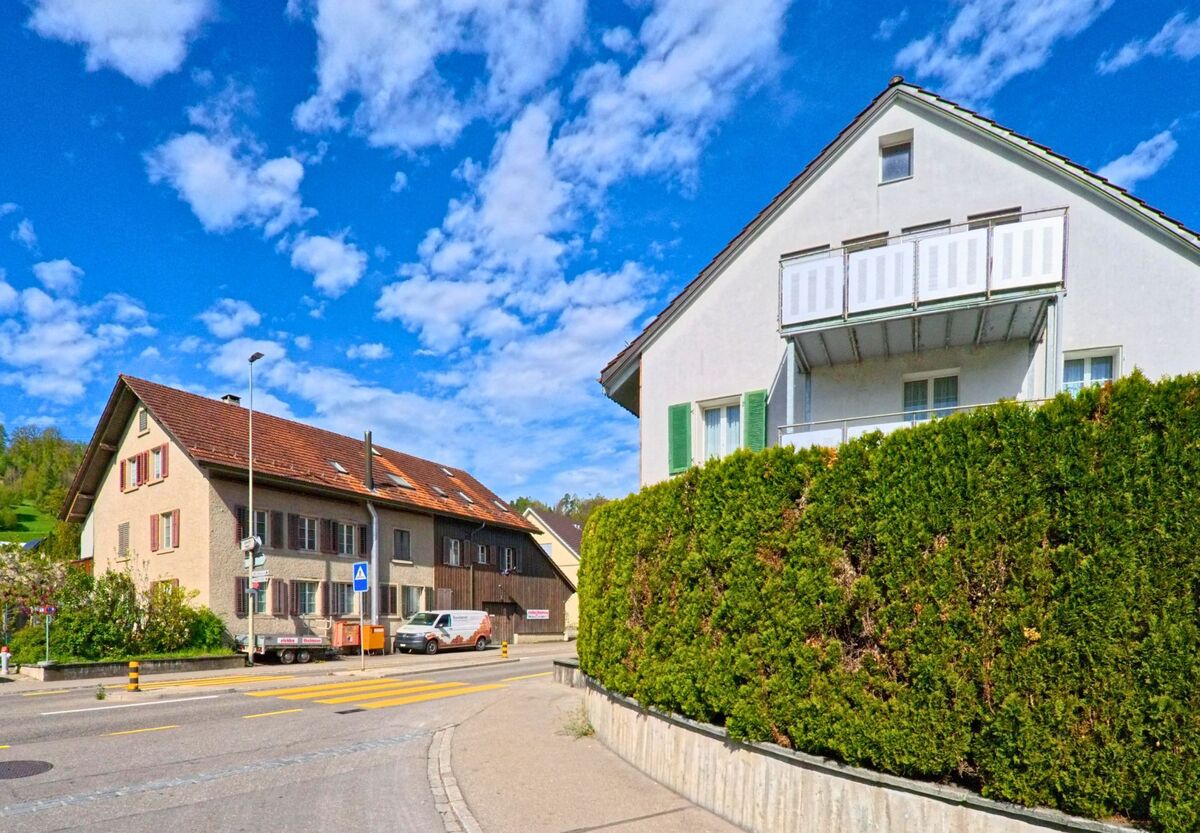 In Rorbas im Kanton Zürich, PLZ 8427, trifft ein schmaler Gehweg auf eine Hauptstrasse mit Fussgängerstreifen und traditionellen Wohnhäusern im Hintergrund.