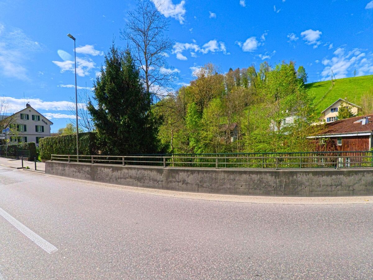 Die Aufnahme zeigt eine Strassenszene in Rorbas im Kanton Zürich, PLZ 8427, mit einem Gehweg, einer Betonmauer und Häusern vor einem bewaldeten Hügel im Hintergrund.