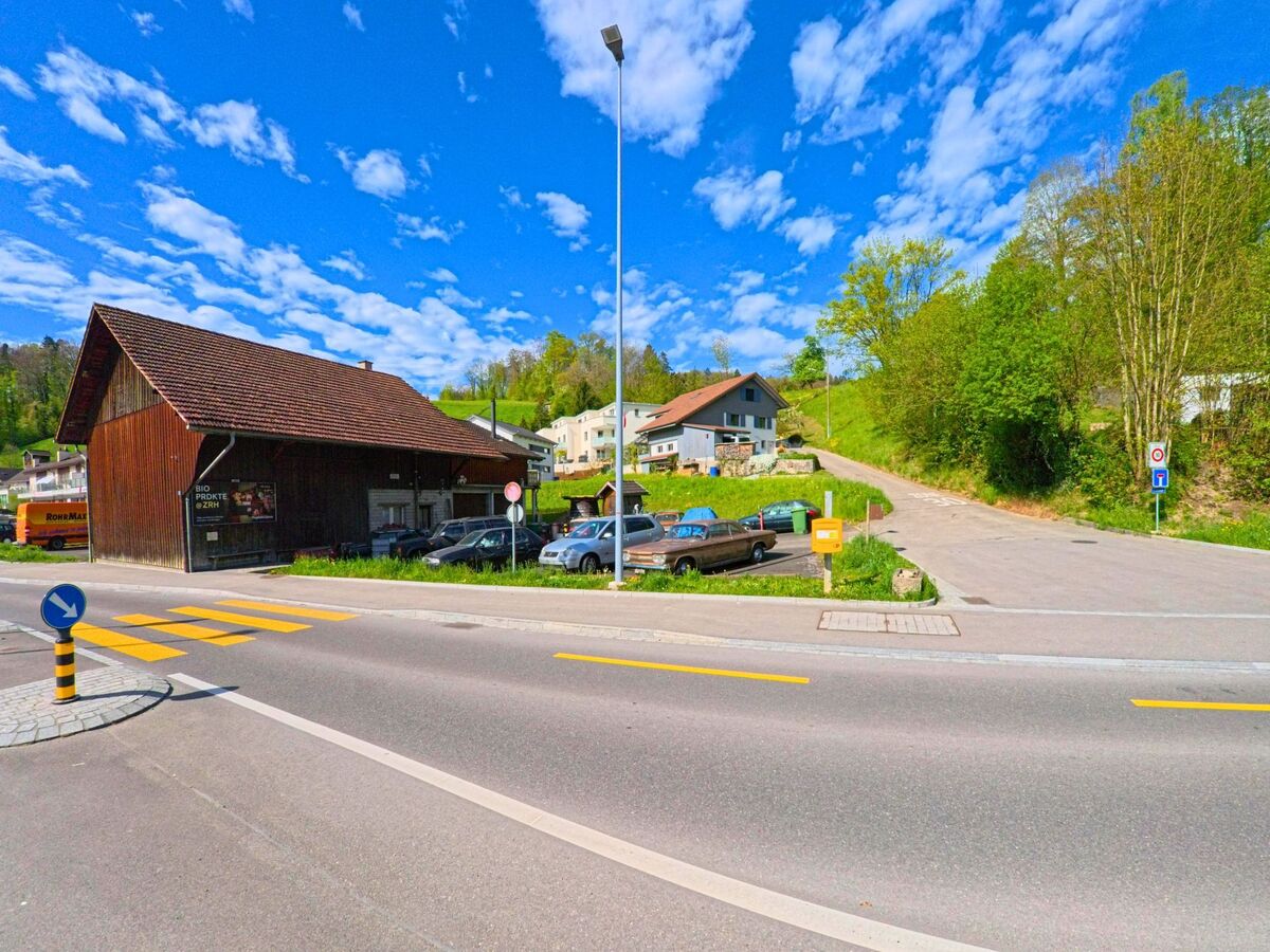 In Rorbas im Kanton Zürich mit der Postleitzahl 8427 steht ein hölzernes Wirtschaftsgebäude an einer Hauptstrasse mit Fussgängerstreifen und angrenzender Sackgasse.