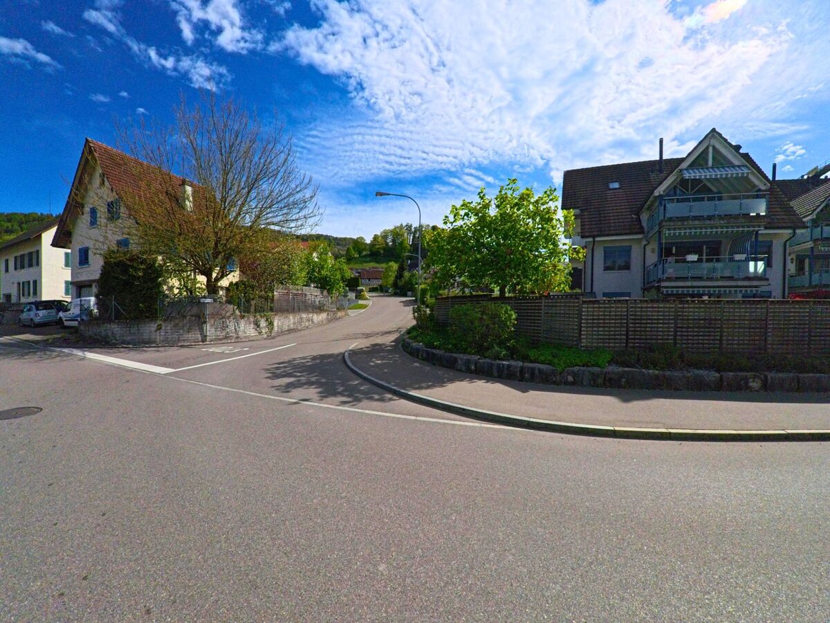 Eine ruhige Seitenstrasse mit Wohnhäusern und Vorgärten verläuft in der Gemeinde Rorbas im Kanton Zürich, PLZ 8427, leicht bergauf in Richtung eines bewaldeten Hangs.