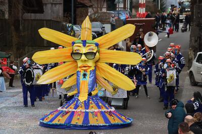 Auf einer Strasse in Fraubrunnen [3317] im Kanton Bern zieht ein farbenfroher Fasnachtsumzug mit grosser Sonnenfigur und Musikgruppe durch den Ort.