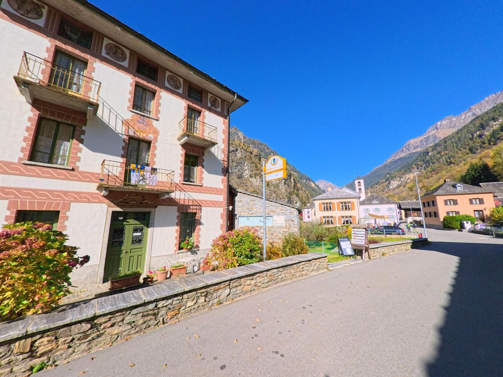 An einer ruhigen Dorfstrasse in Rossa (6548, Kanton Graubünden) steht ein historisches Wohnhaus mit Balkonen vor alpiner Bergkulisse.