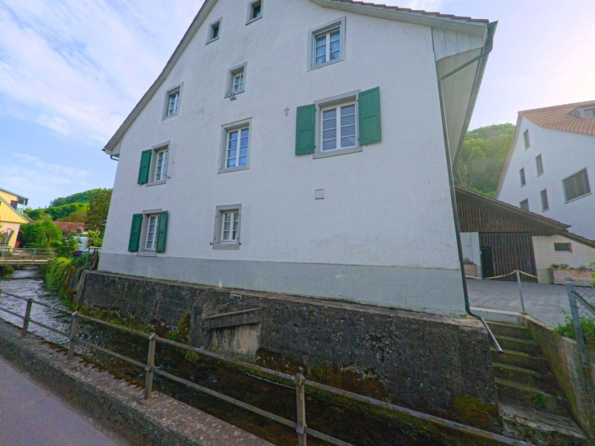 In Rothenfluh (4467, Kanton Baselland) fliesst ein schmaler Bach direkt an einem weissen Haus mit gruenen Fensterlaeden vorbei, umgeben von weiteren Gebaeuden und Huegeln.