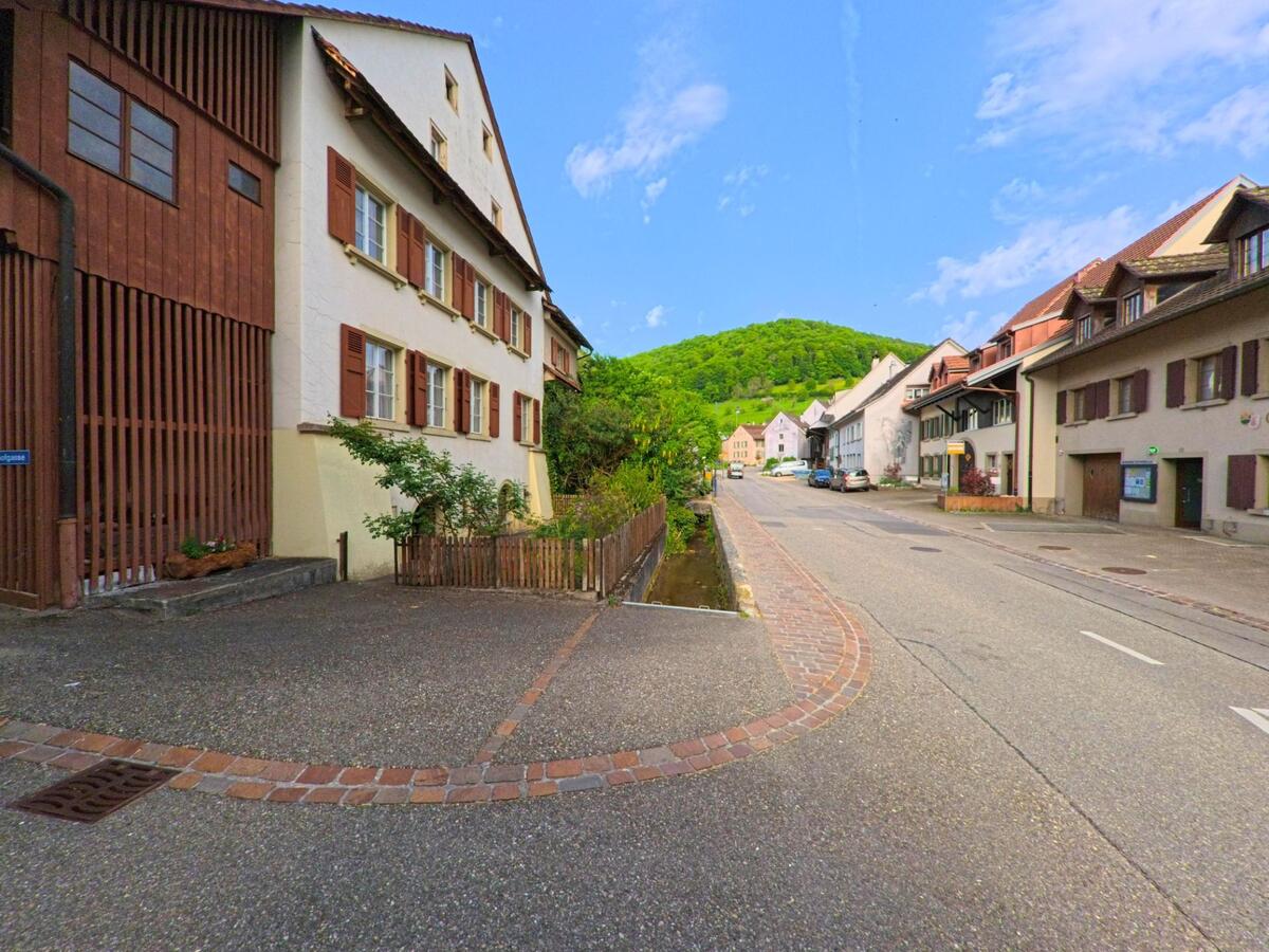 Entlang einer gepflasterten Strasse in Rothenfluh (4467, Kanton Baselland) verlaeuft ein schmaler Wasserlauf, gesaeumt von historischen Haeusern und gruener Huegellandschaft im Hintergrund.