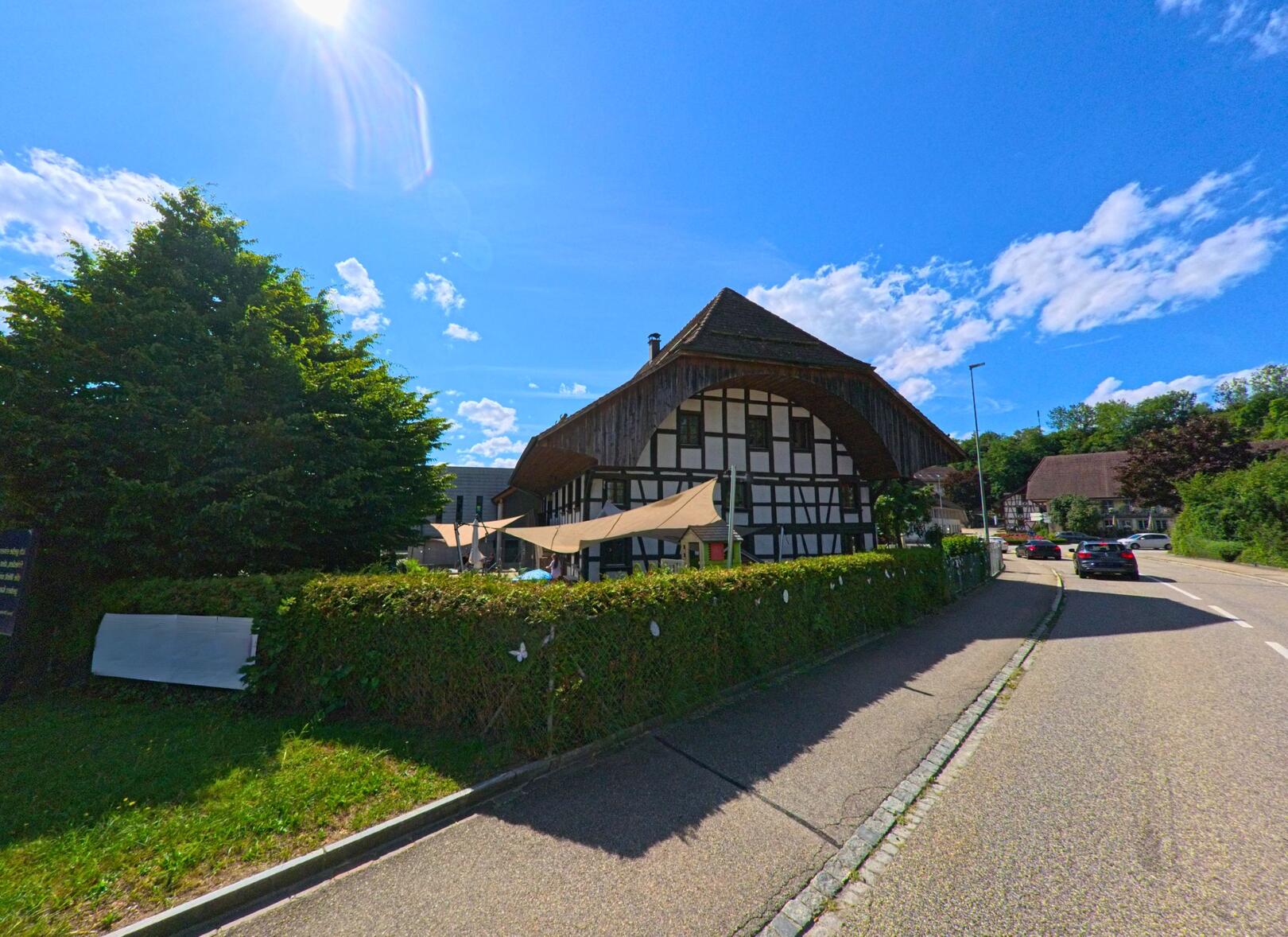 In Rothrist (4852, Kanton Aargau) steht ein grosses Fachwerkhaus mit weit ausladendem Dach direkt an einer leicht geschwungenen Strasse bei strahlendem Sonnenschein.
