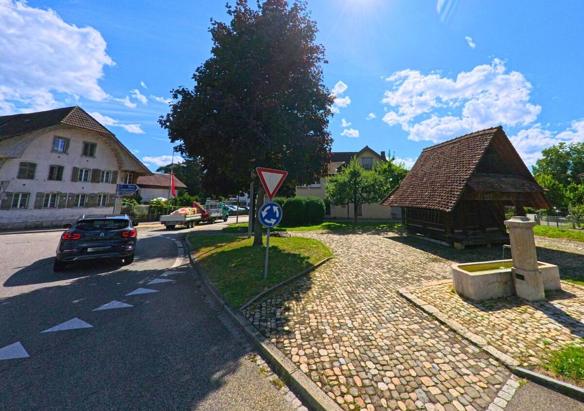 In einem kleinen Kreisverkehr in Rothrist (4852, Kanton Aargau) befinden sich ein Brunnen und ein traditionelles Holzgebaeude auf gepflastertem Platz, umgeben von alten Wohnhaeusern.