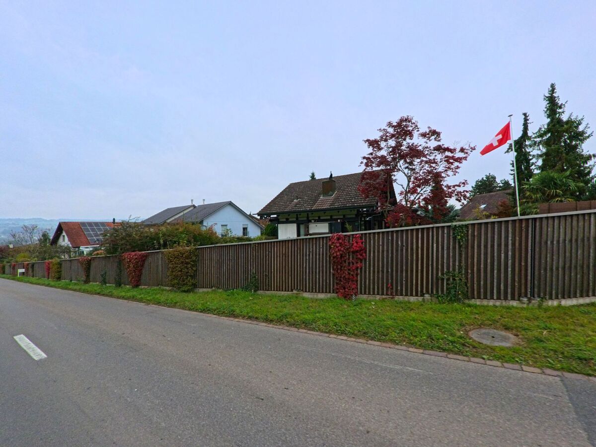 Entlang einer ruhigen Strasse in Rottenschwil (8919, Kanton Aargau) stehen Einfamilienhaeuser hinter einem langen Holzzaun, vor dem eine Schweizer Fahne im Wind weht.