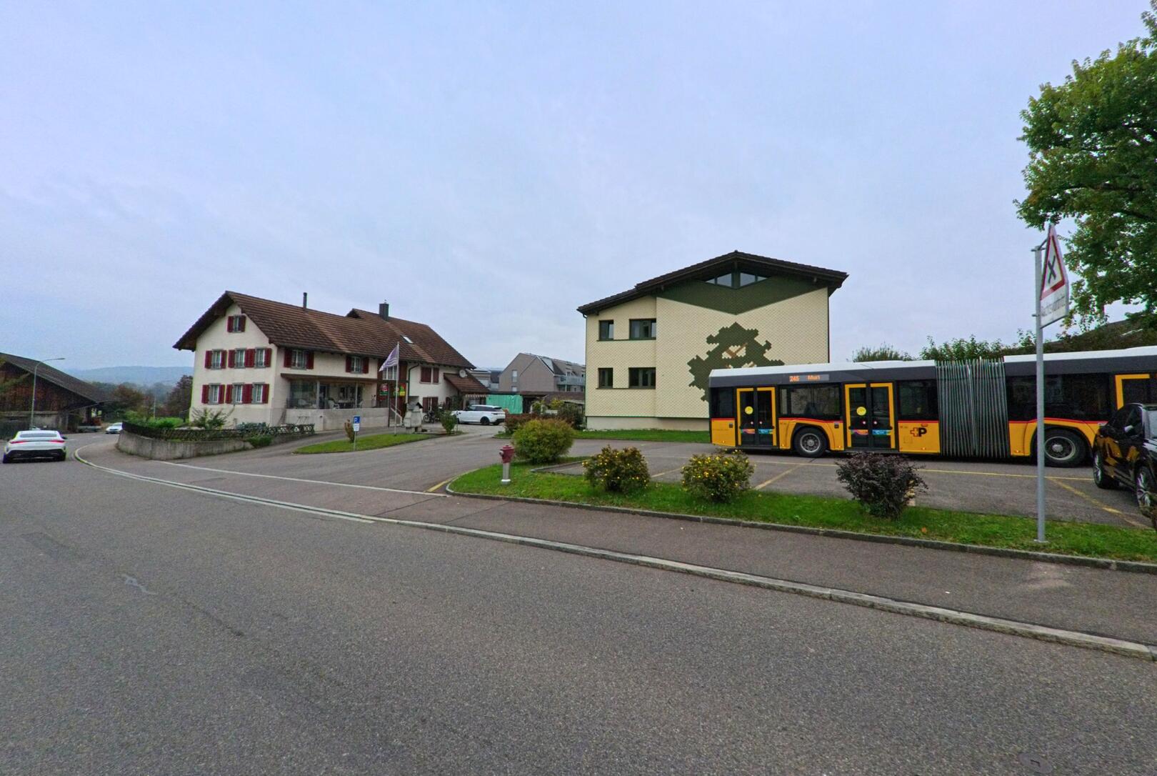 Ein Postauto haelt an der Hauptstrasse von Rottenschwil (8919, Kanton Aargau) vor einem hellen Wohnhaus mit rotem Fensterladen und einem modernen Gebaeude mit Wandmosaik.