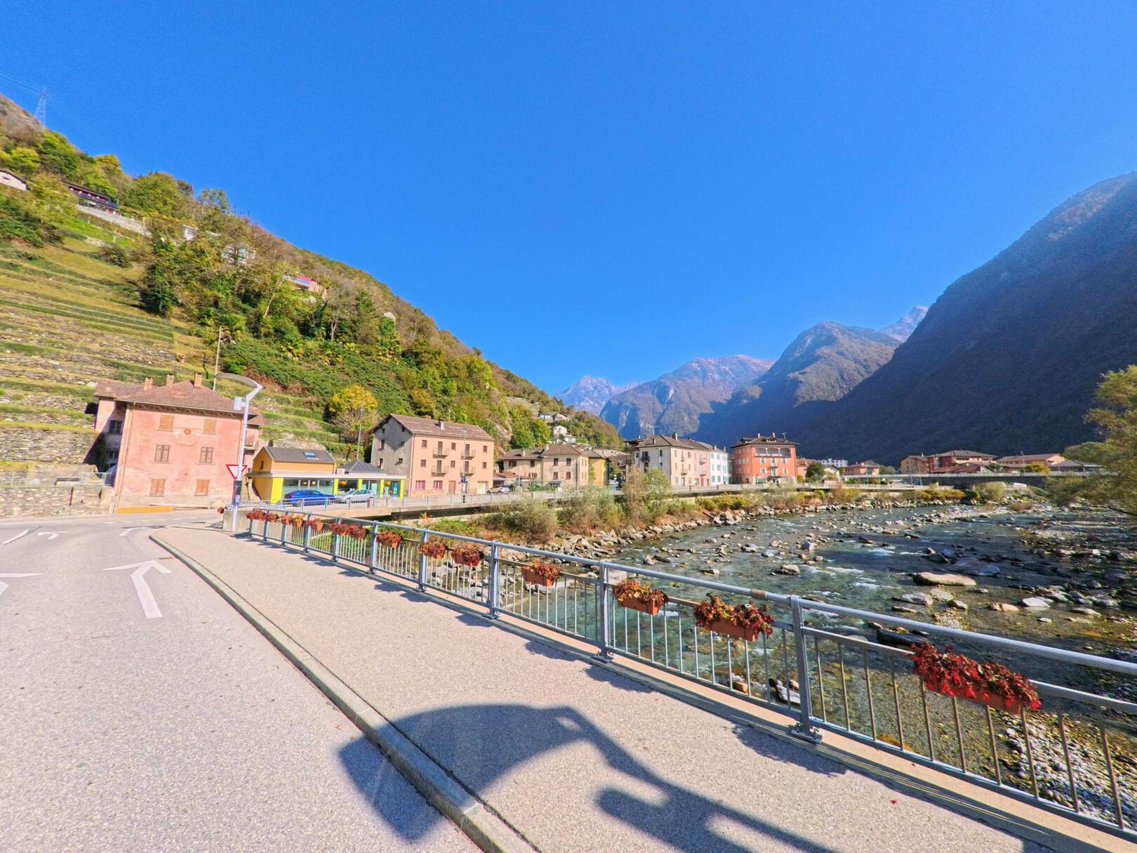 Entlang einer Brücke in Roveredo (6535, Kanton Graubünden) verläuft ein Gehweg mit Blick auf den Fluss, farbige Wohnhäuser und die umliegende Berglandschaft.