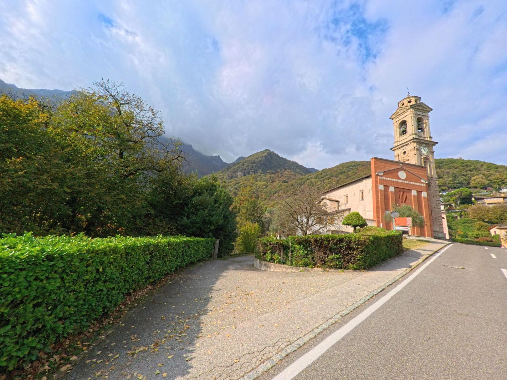 Ein schmaler Gehweg führt in Rovio (6821, Kanton Tessin) an einer Kirche mit markantem Glockenturm vorbei, eingerahmt von Hecken und bewaldeten Berghängen.