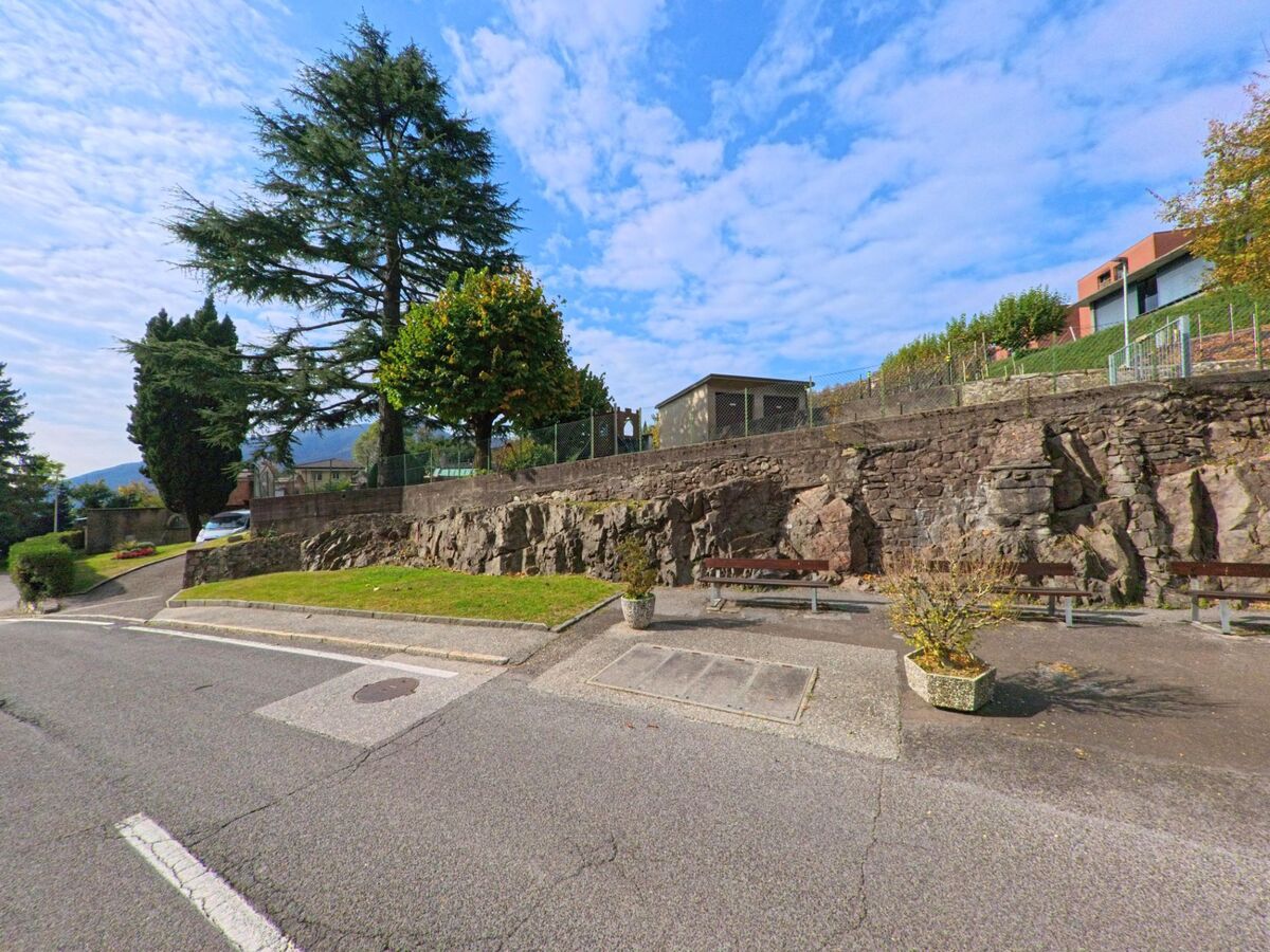 Eine kleine Sitzgelegenheit mit Bänken liegt in Rovio (6821, Kanton Tessin) am Rand einer Strasse vor einer Natursteinmauer und hohen Bäumen.