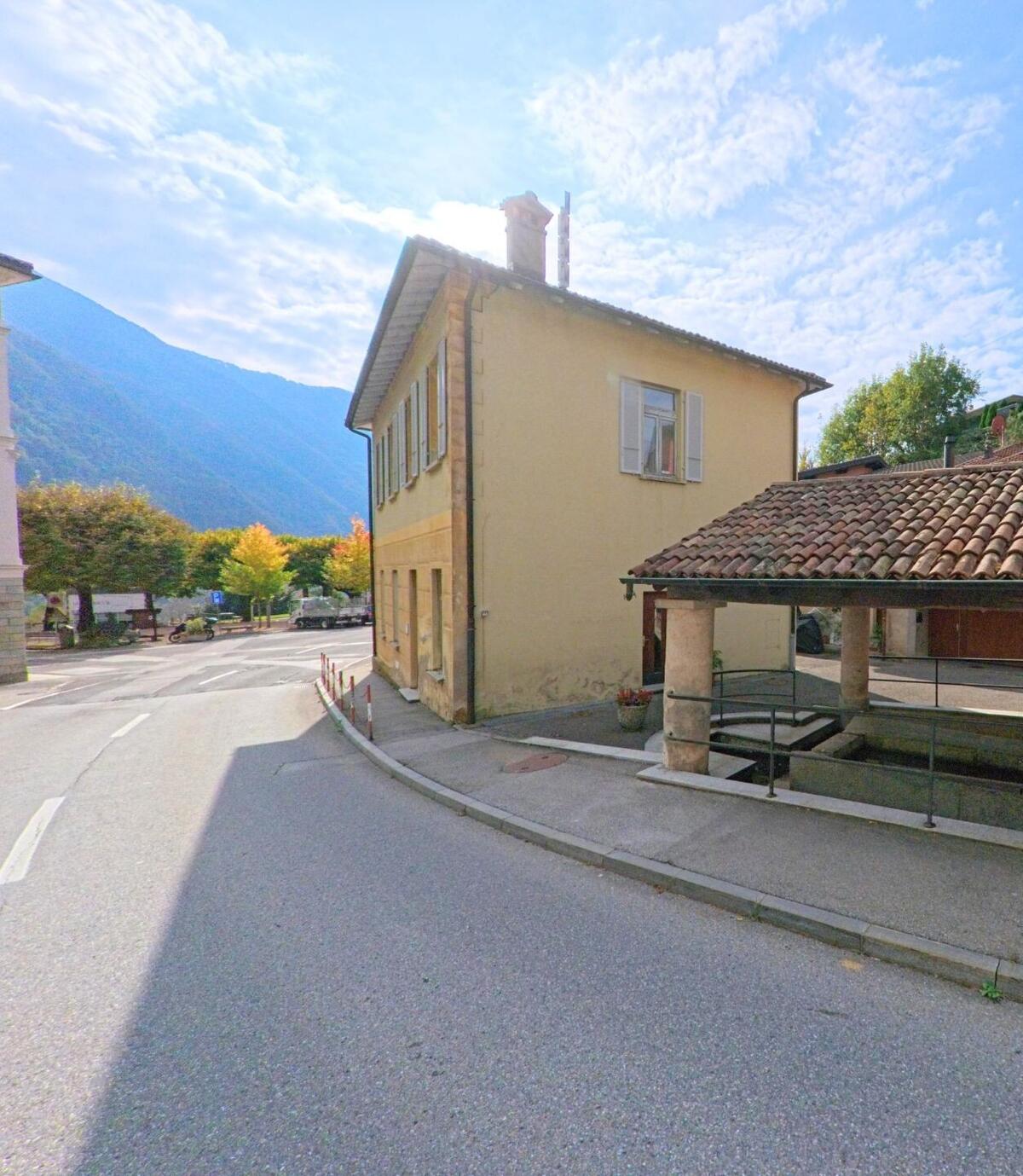 An einer ruhigen Dorfstrasse in Rovio (6821, Kanton Tessin) steht ein helles Wohnhaus neben einem überdachten Brunnen vor bewaldeten Berghängen.