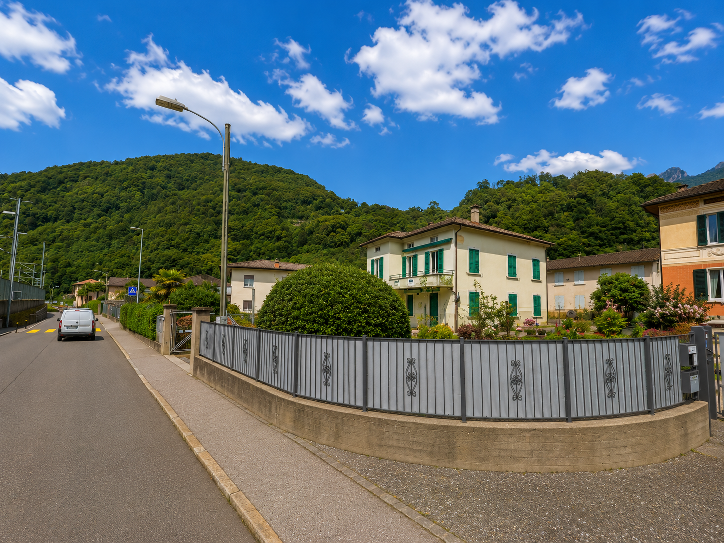 Entlang einer ruhigen Wohnstrasse in Rovio (6821, Kanton Tessin) stehen helle Häuser mit grünen Fensterläden vor dicht bewaldeten Hängen.