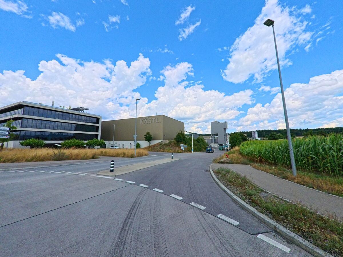 In Rubigen (3113, Kanton Bern) führt eine breite Strasse durch das Industriegebiet mit modernen Gebäuden, darunter ein markanter Bau mit der Aufschrift „Kästli“, angrenzend an ein Maisfeld.