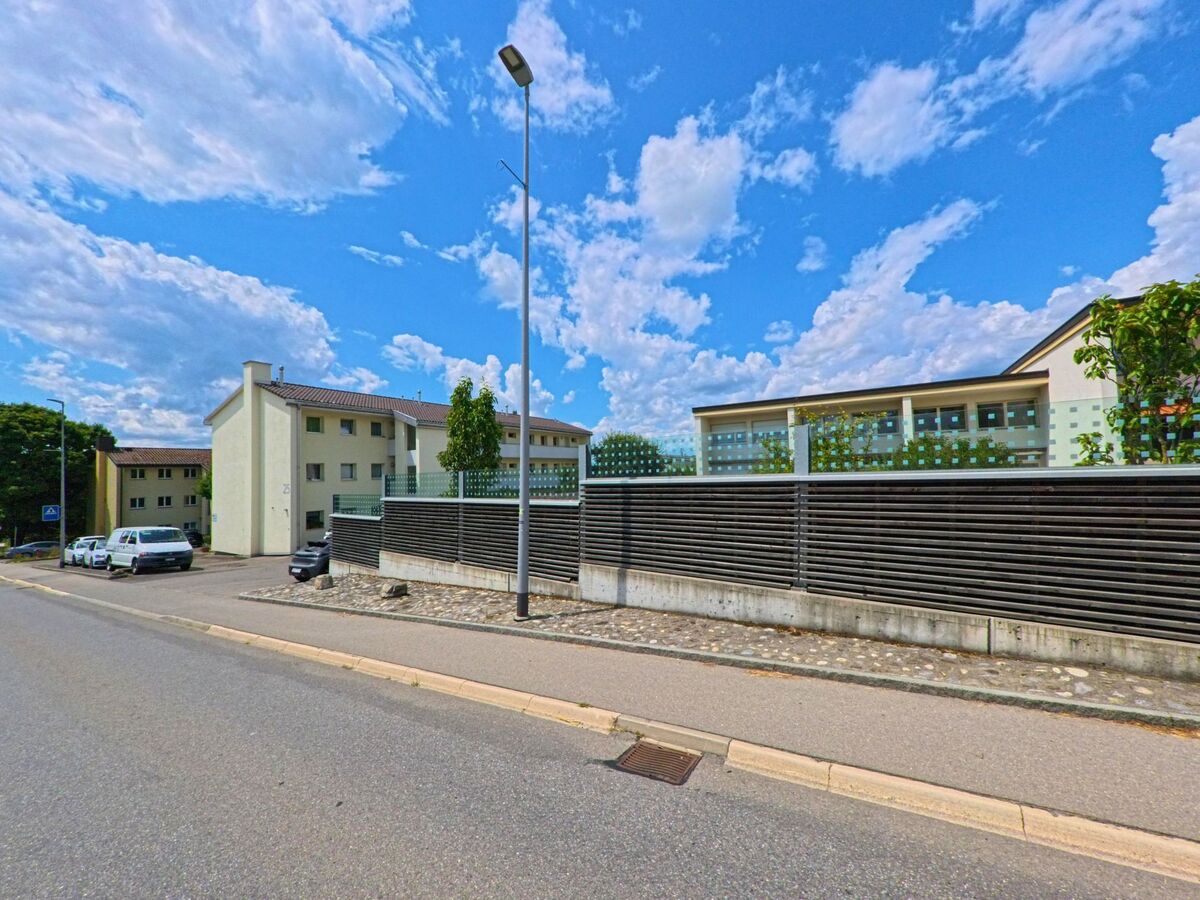 In Rubigen (3113, Kanton Bern) stehen mehrere moderne Wohngebäude mit hellen Fassaden entlang einer ruhigen Quartierstrasse, begrenzt durch eine Sichtschutzwand mit Glas- und Holz­elementen.