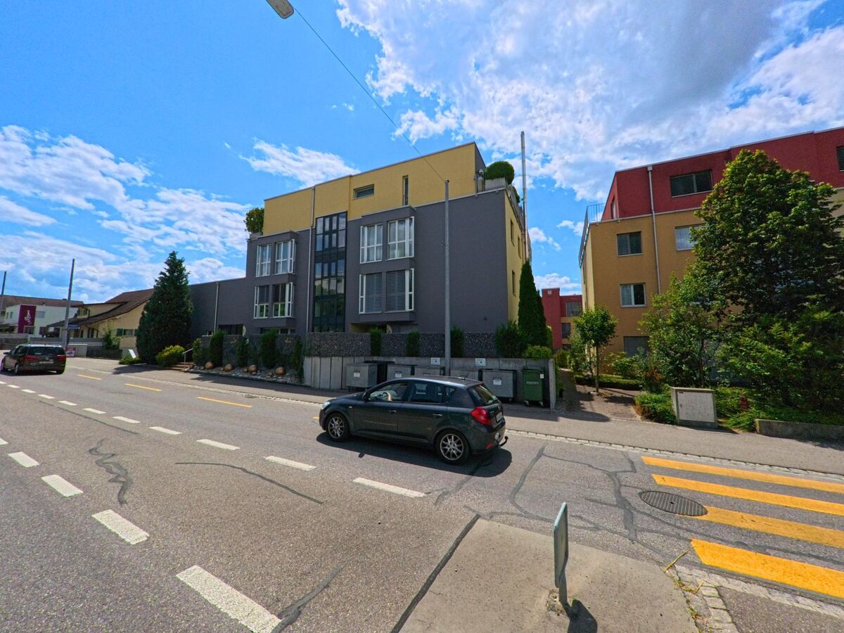 In Rubigen (3113, Kanton Bern) stehen moderne, farblich kontrastierende Wohnhäuser mit grossen Fenstern entlang einer Hauptstrasse mit markiertem Fussgängerstreifen.