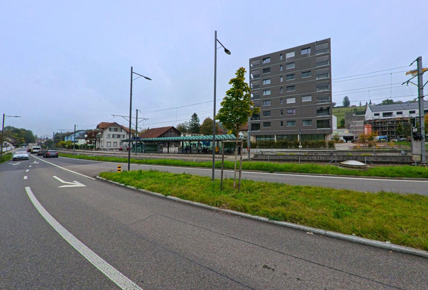 Am Bahnhof von Rudolfstetten-Friedlisberg (8964, Kanton Aargau) verlaufen Strasse und Bahnlinie nebeneinander, flankiert von modernen und traditionellen Gebäuden.