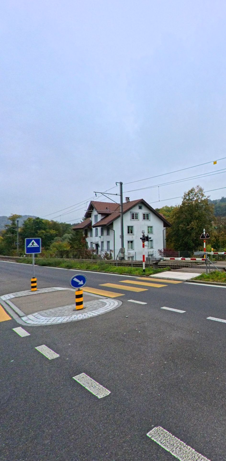 Ein Bahnübergang mit Zebrastreifen und einem weissen Wohnhaus im Hintergrund befindet sich in Rudolfstetten-Friedlisberg (8964, Kanton Aargau).