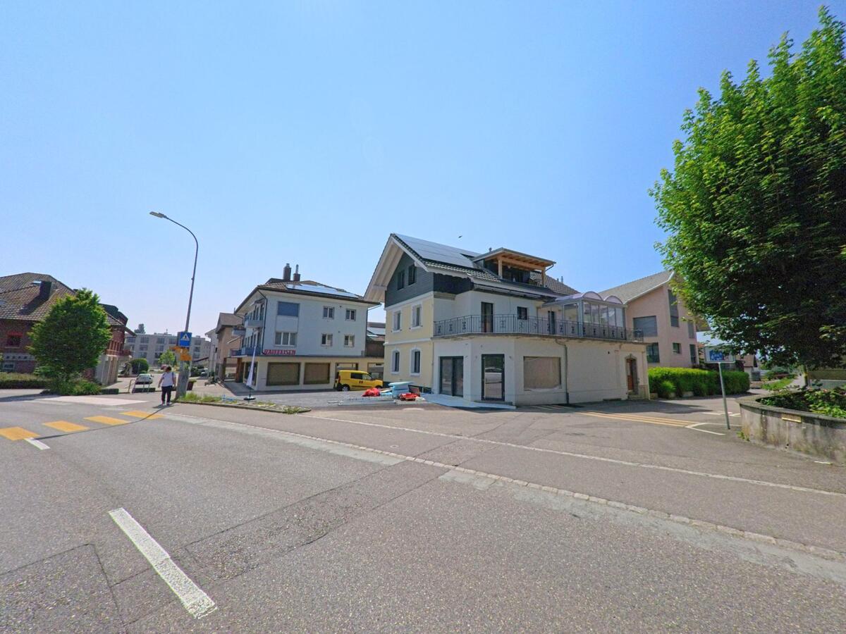 Ein helles Wohn- und Geschäftshaus steht an einer Kreuzung in Rüdtligen-Alchenflüh (3422, Kanton Bern) bei klarem Sommerwetter.