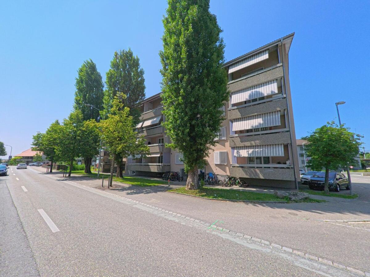 Mehrstöckige Wohnhäuser mit Balkonen und schattenspendenden Bäumen stehen in Rüdtligen-Alchenflüh (3422, Kanton Bern) an einer breiten, sonnigen Strasse.