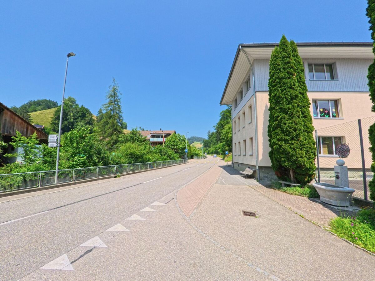 In Rüegsau (3418, Kanton Bern) führt eine ruhige Straße an gepflegten Gebäuden und üppigem Grün entlang, mit einem klaren blauen Himmel im Hintergrund.