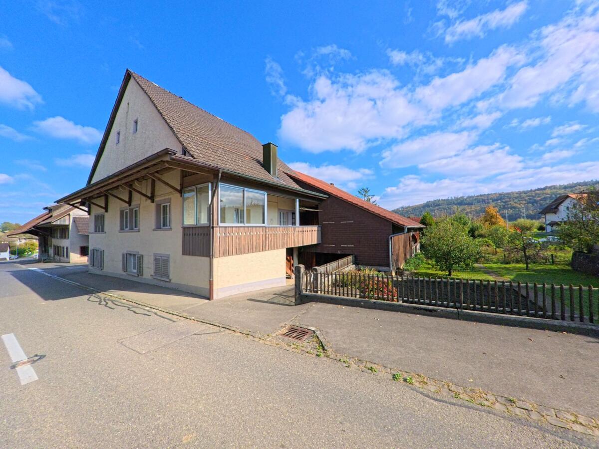 Ein traditionelles Bauernhaus mit grossem Scheunentrakt steht in Rüfenach (5235, Kanton Aargau) an einer ruhigen Strasse mit Blick auf Wiesen und Hügel.