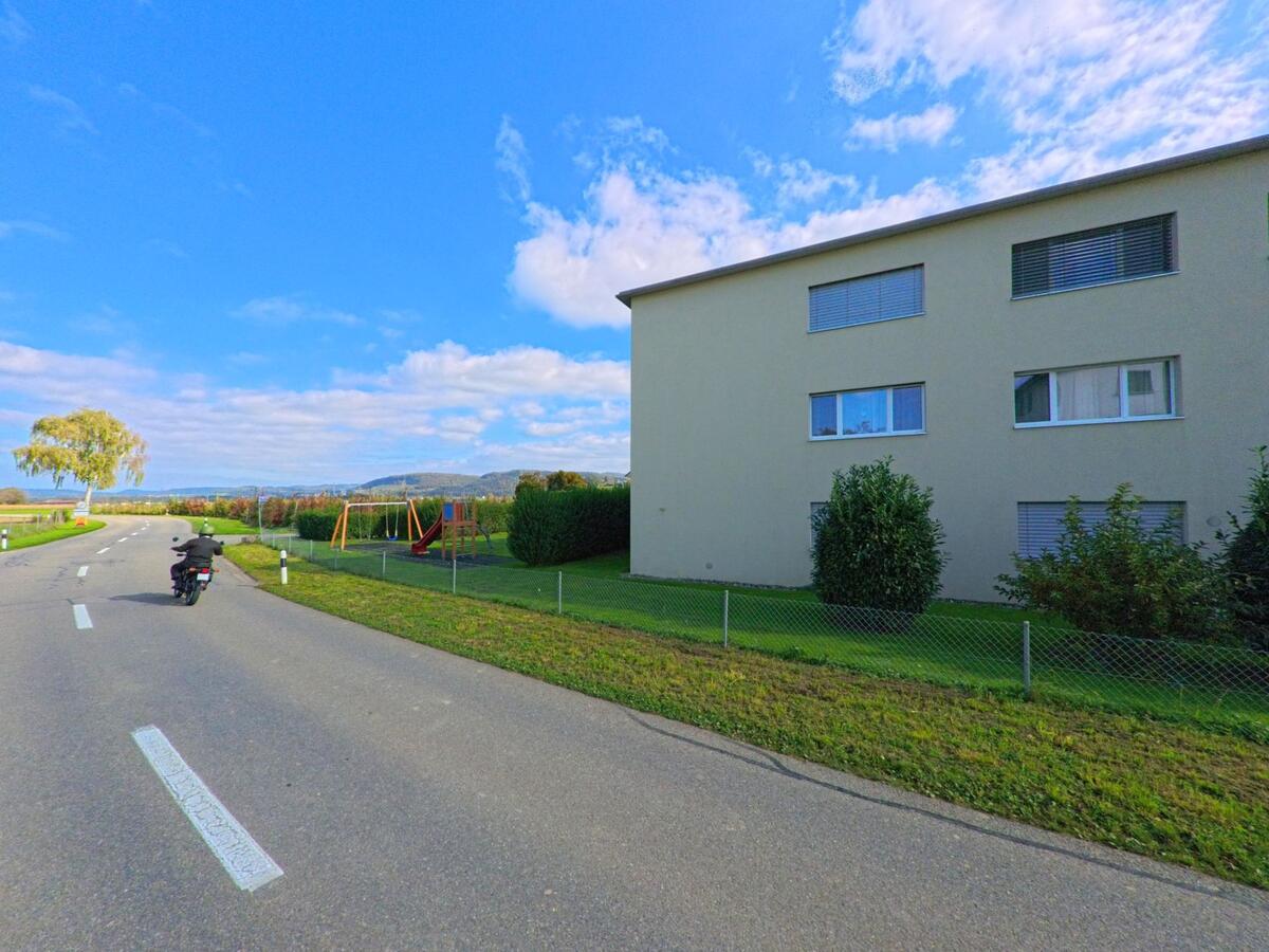 Ein Motorradfahrer fährt in Rüfenach (5235, Kanton Aargau) an einem modernen Wohnhaus mit angrenzendem Spielplatz und weiter Sicht über die Landschaft vorbei.
