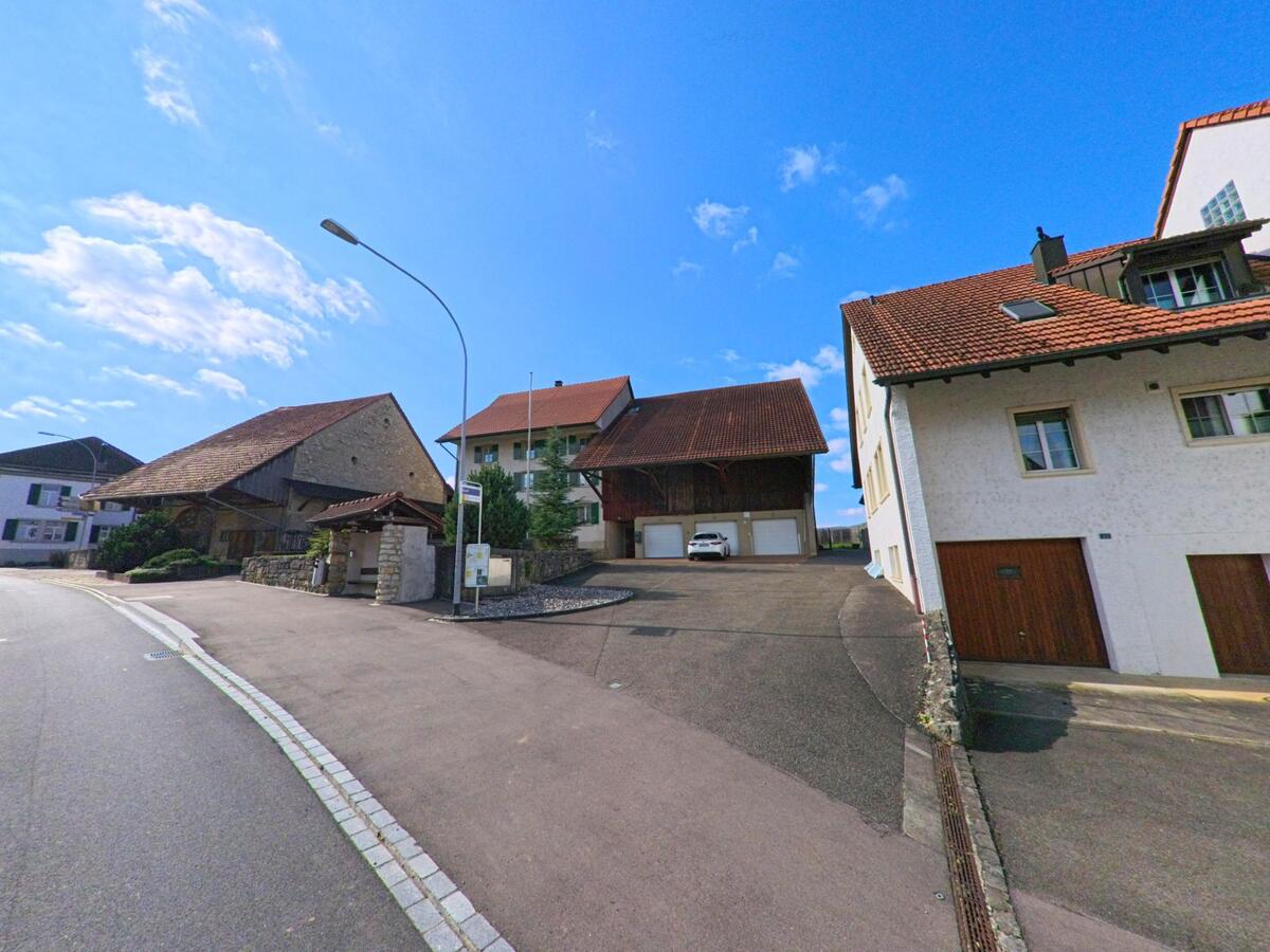 Mehrere gepflegte Wohnhäuser und Scheunen mit roten Ziegeldächern stehen an einer leicht ansteigenden Strasse in Rüfenach (5235, Kanton Aargau).
