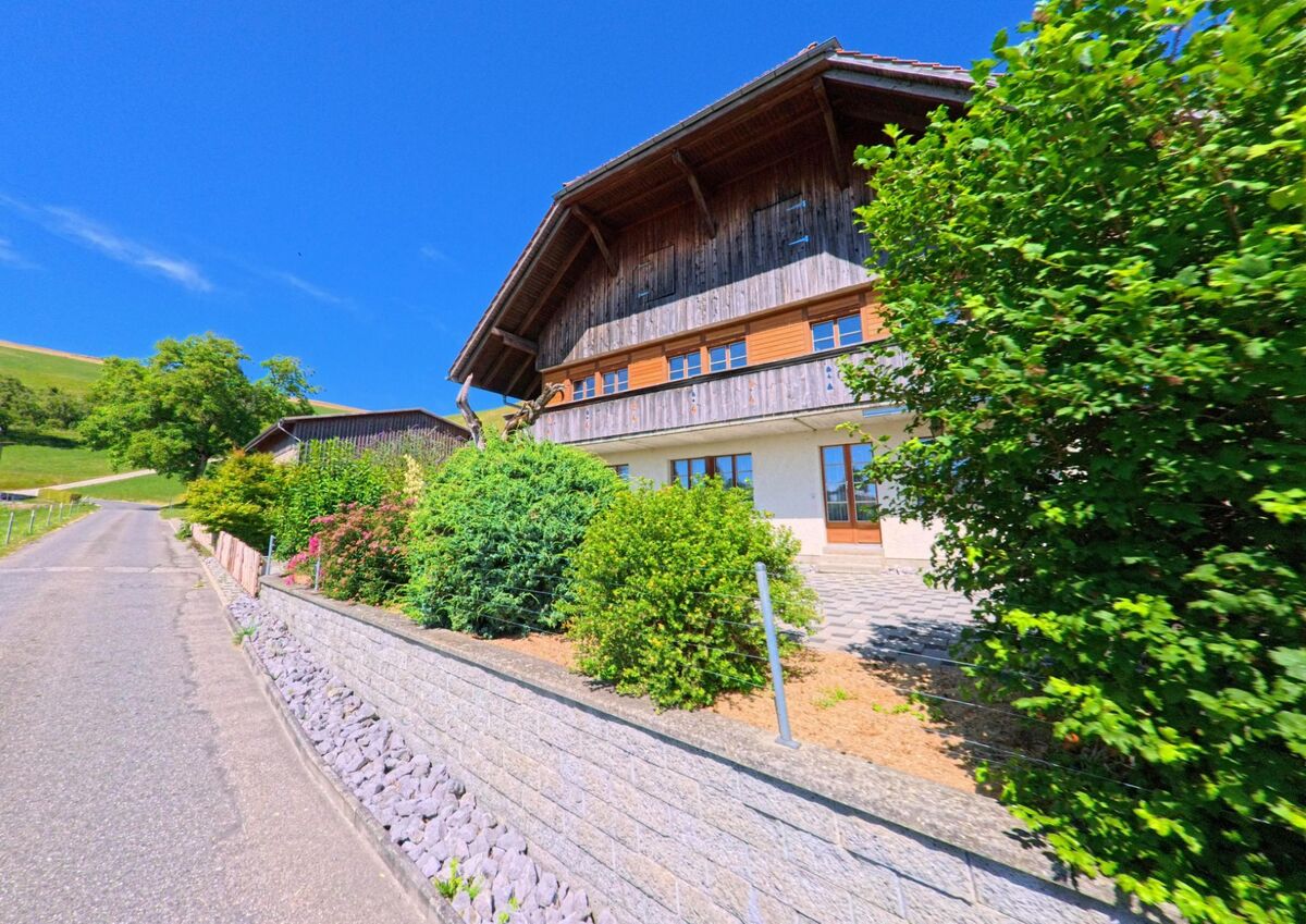 In Rüeggisberg (3089, Kanton Bern) steht ein modernes Bauernhaus mit Holzfassade und gepflegtem Vorgarten an einem sanft ansteigenden Strassenabschnitt mit Blick auf die umliegenden Felder.