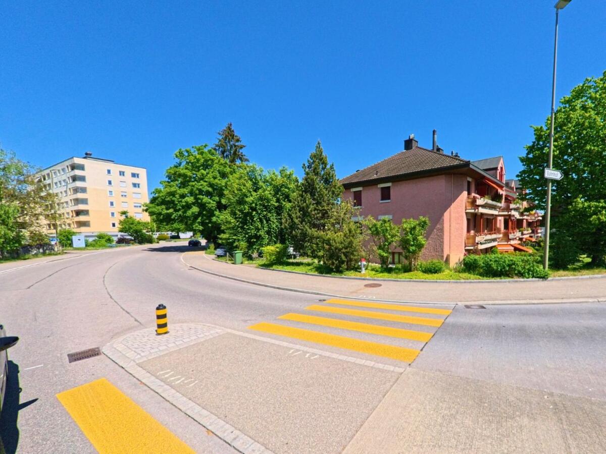 Ein Fussgängerstreifen führt über eine Kurve in einem Wohngebiet von Rümlang (8153, Kanton Zürich), flankiert von mehrstöckigen Wohnhäusern und dichtem Grün.