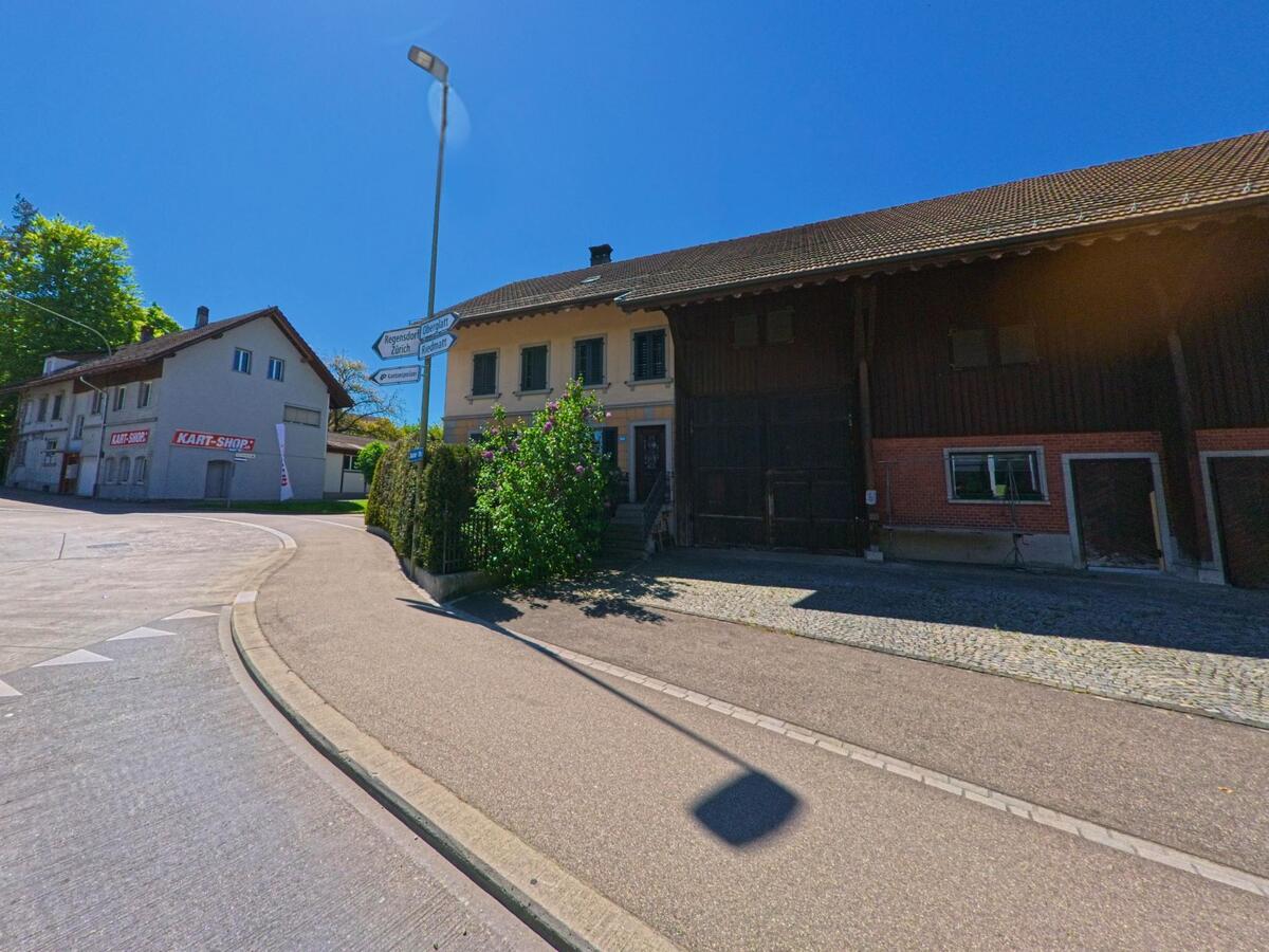 In Rümlang (8153, Kanton Zürich) treffen ein traditioneller Bauernhof und ein Kart-Shop an einer Strassengabelung mit Wegweisern unter klarem Himmel aufeinander.